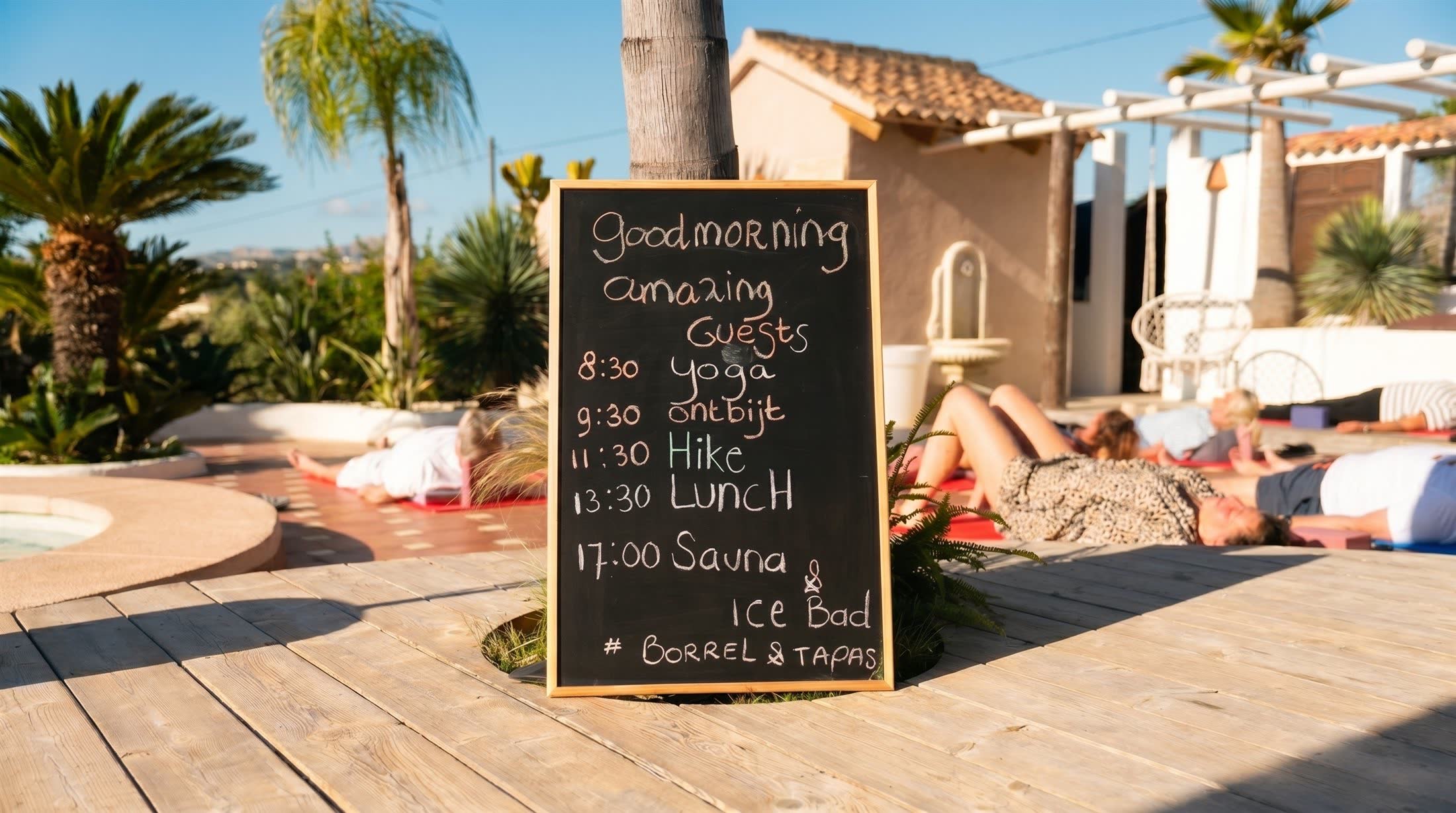 Krijtbord bij het zwembad met het handgeschreven dagprogramma van een retreat — yoga, ontbijt, hike, lunch, sauna en ijsbad — terwijl gasten ontspannen in de ochtendzon