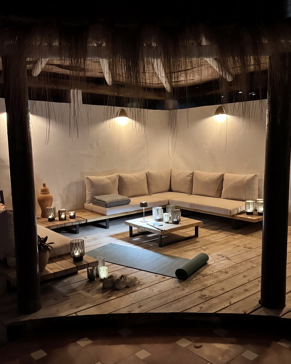 De lounge onder de rieten palapa bij avond: kaarslantaarns, warme wandlampen, lage banken en een uitgerolde yogamat op het houten dek