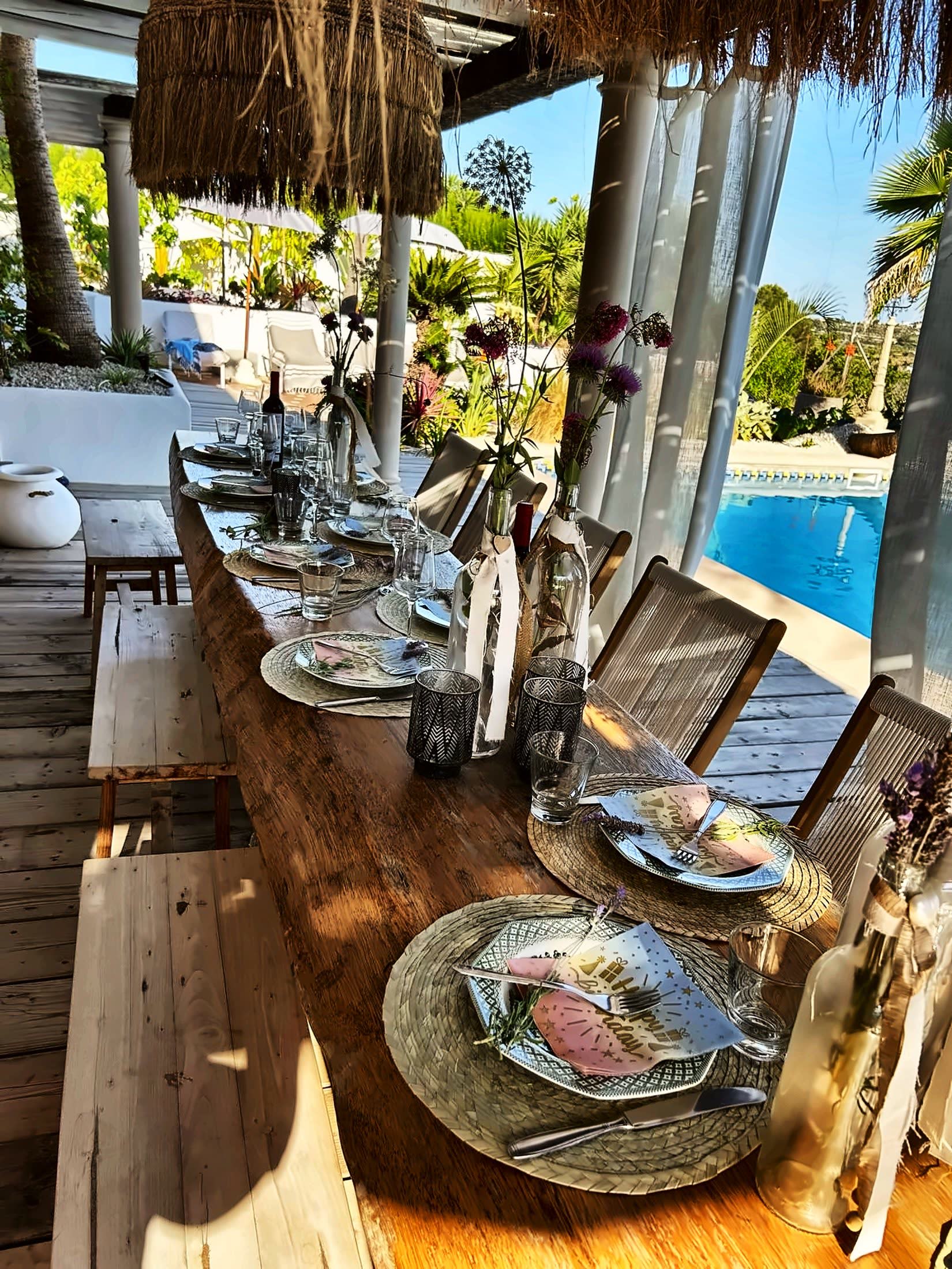 Gedekte lange tafel met wildbloemen onder de pergola, met het azuurblauwe zwembad ernaast